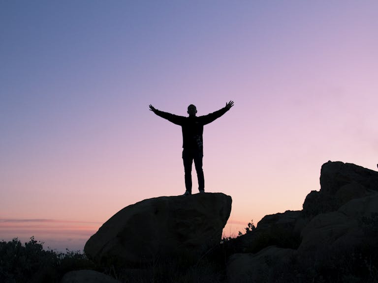 VirtusPlays Silhouette of a man on a rock with arms raised, embracing the sunset sky.