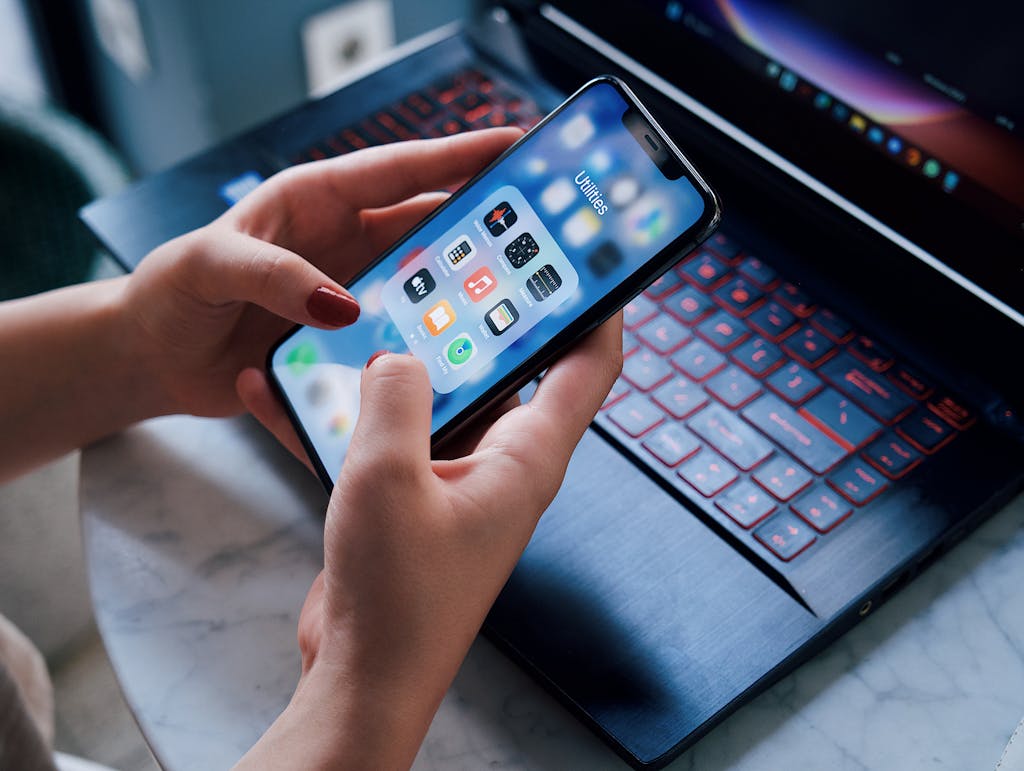 uitly.blog Close-up of a woman holding a smartphone showing utilities apps while sitting by a laptop indoors.