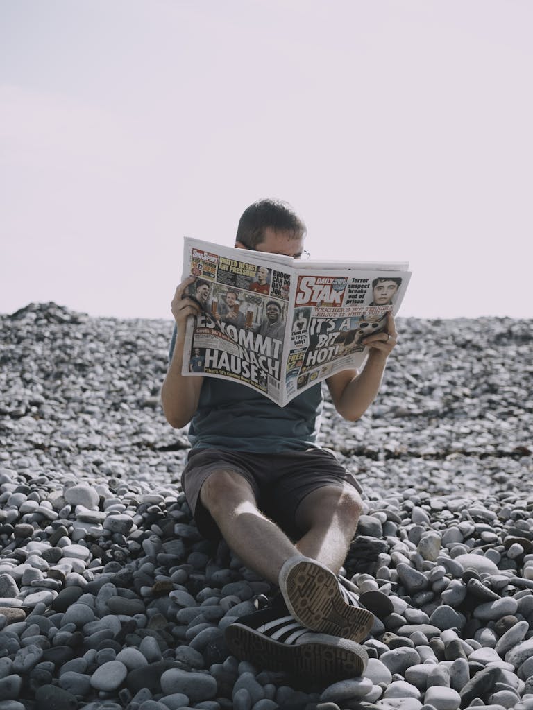 Newstook Man enjoying leisure time reading a newspaper on a stony beach under bright daylight.