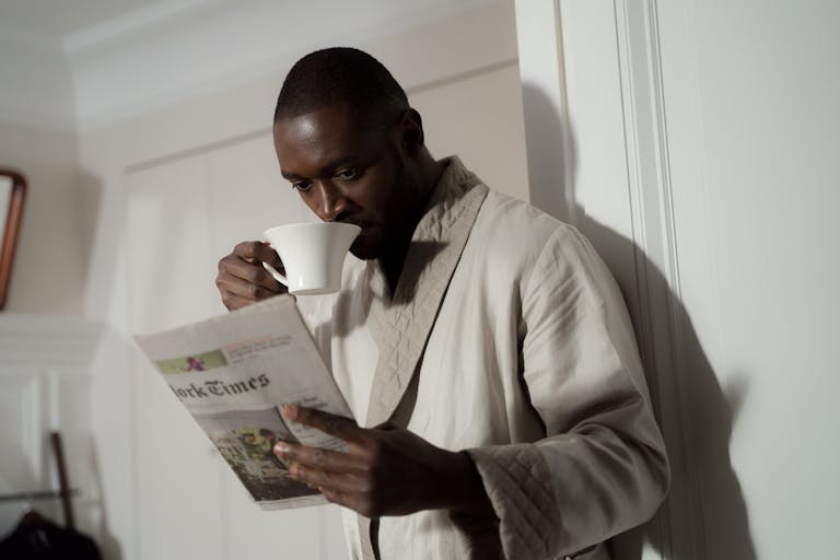 Newstook Man in a bathrobe enjoys coffee and reads a newspaper indoors.