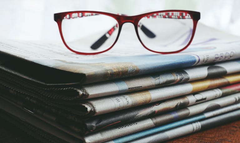 ventsmagazine.cloud Red eyeglasses resting on a stack of newspapers symbolize reading and information.