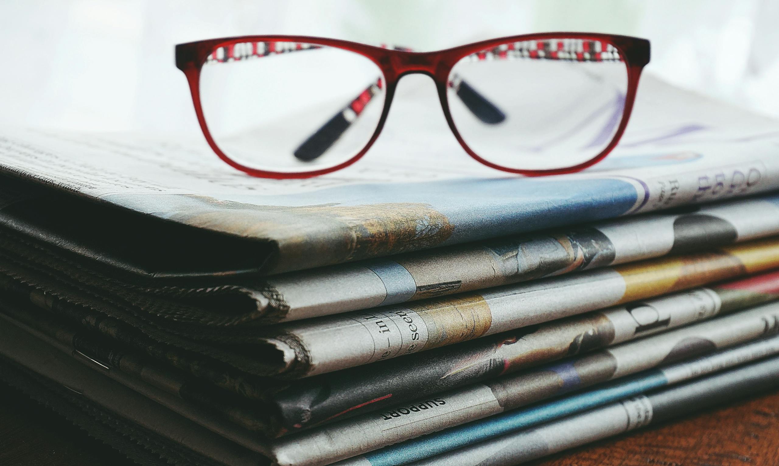 ventsmagazine.cloud Red eyeglasses resting on a stack of newspapers symbolize reading and information.