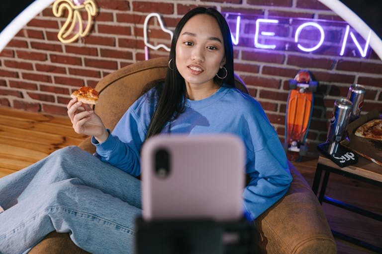 FamousParentingMomlife Young Asian woman streaming video while enjoying pizza in a cozy setup with neon lights.